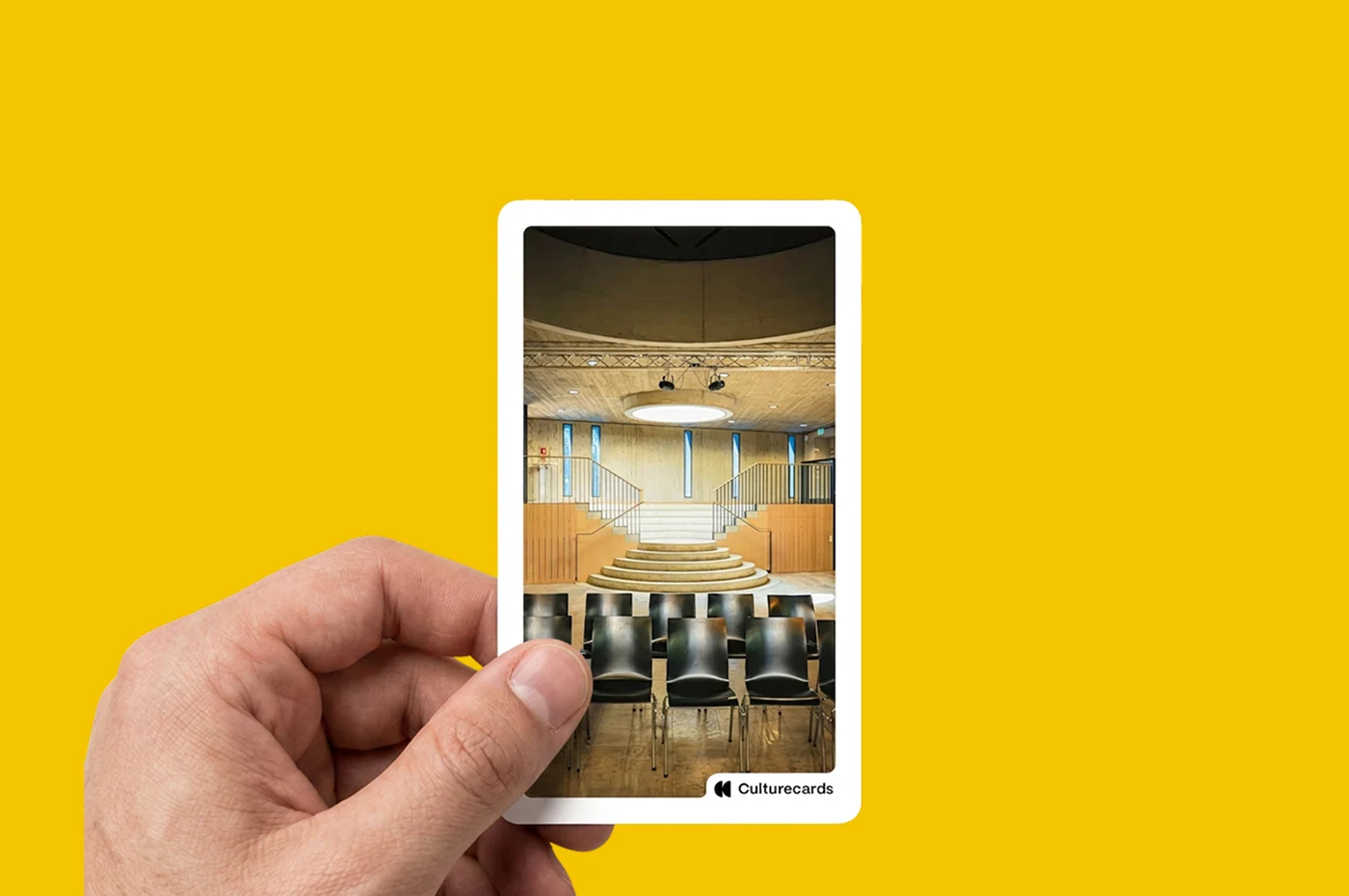 Eine Hand hält eine Culturecards-Karte mit dem Foto eines Veranstaltungssaals mit Stufenbühne und Stuhlreihen.